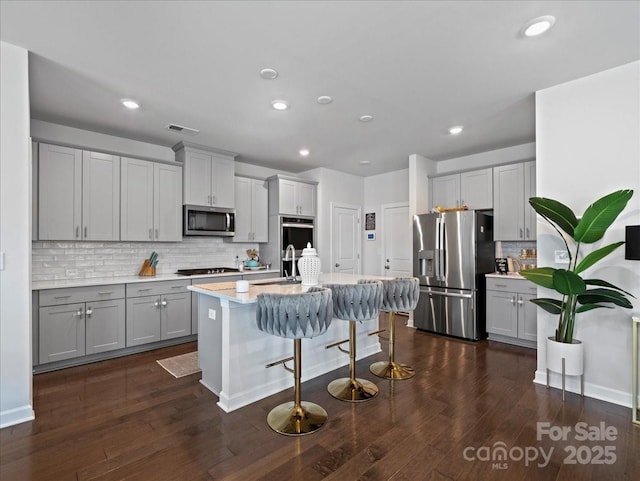 kitchen featuring dark wood-type flooring, a breakfast bar area, appliances with stainless steel finishes, an island with sink, and backsplash