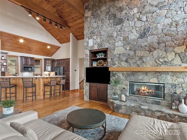 living room featuring rail lighting, high vaulted ceiling, light hardwood / wood-style floors, a stone fireplace, and wooden ceiling