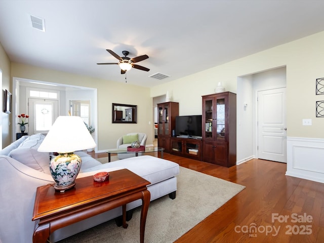 living room with dark wood-style floors, wainscoting, visible vents, and ceiling fan