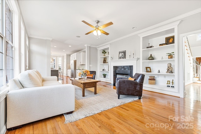 living room with crown molding, a tile fireplace, ceiling fan, light hardwood / wood-style floors, and built in shelves