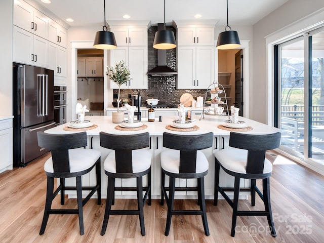 kitchen featuring high end refrigerator, a center island with sink, light wood-type flooring, pendant lighting, and white cabinets