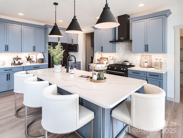 kitchen featuring decorative light fixtures, decorative backsplash, a kitchen island with sink, black appliances, and wall chimney exhaust hood