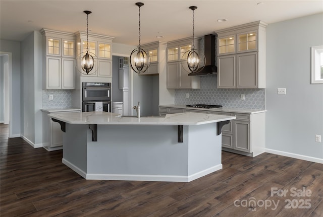 kitchen featuring double oven, an island with sink, sink, light stone countertops, and wall chimney range hood