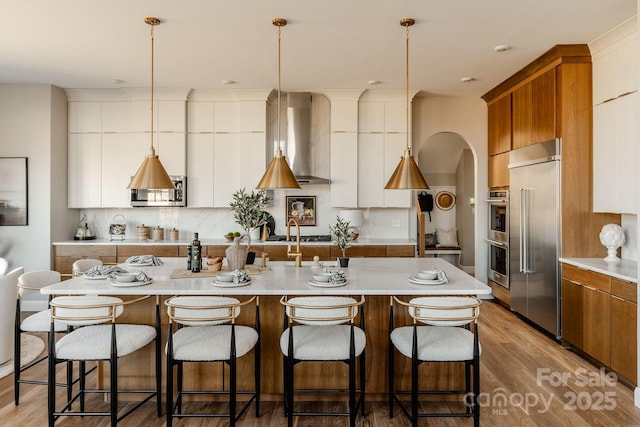 kitchen featuring pendant lighting, wall chimney range hood, a kitchen island with sink, stainless steel appliances, and white cabinets