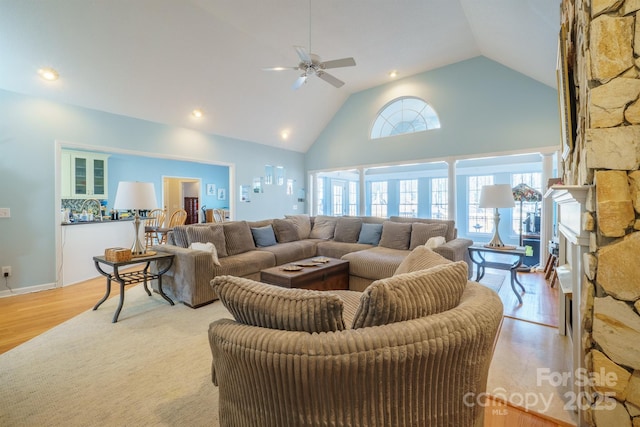 living room featuring a stone fireplace, high vaulted ceiling, ceiling fan, and light hardwood / wood-style flooring