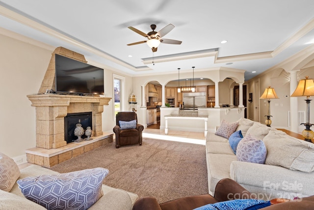 living area with light carpet, ornamental molding, a tiled fireplace, a tray ceiling, and arched walkways