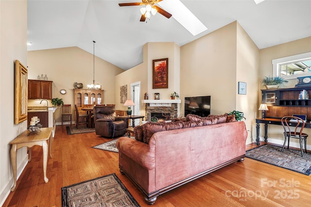 living room with wood-type flooring, lofted ceiling with skylight, ceiling fan with notable chandelier, and a stone fireplace