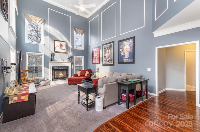 carpeted living room featuring crown molding, a towering ceiling, ceiling fan, and a high end fireplace