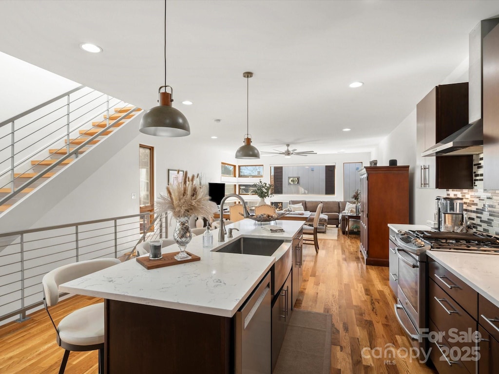 kitchen featuring appliances with stainless steel finishes, pendant lighting, an island with sink, sink, and wall chimney range hood