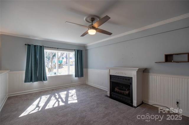 unfurnished living room featuring a wainscoted wall, a fireplace with flush hearth, carpet flooring, crown molding, and ceiling fan