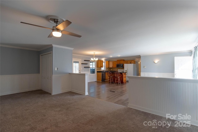 unfurnished living room with ceiling fan with notable chandelier, ornamental molding, carpet, and wainscoting