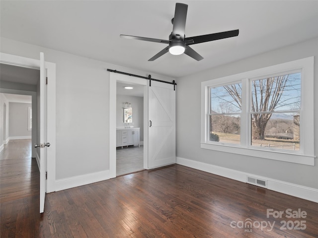 unfurnished bedroom featuring a barn door, dark hardwood / wood-style floors, ceiling fan, and ensuite bath