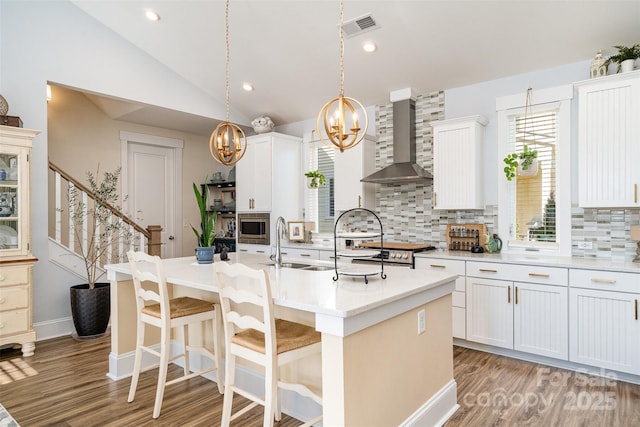 kitchen featuring hanging light fixtures, appliances with stainless steel finishes, an island with sink, wall chimney range hood, and white cabinets