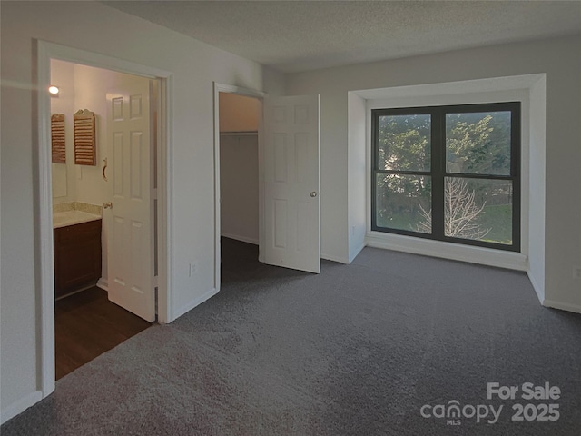 unfurnished bedroom with dark colored carpet, a walk in closet, ensuite bathroom, and a textured ceiling