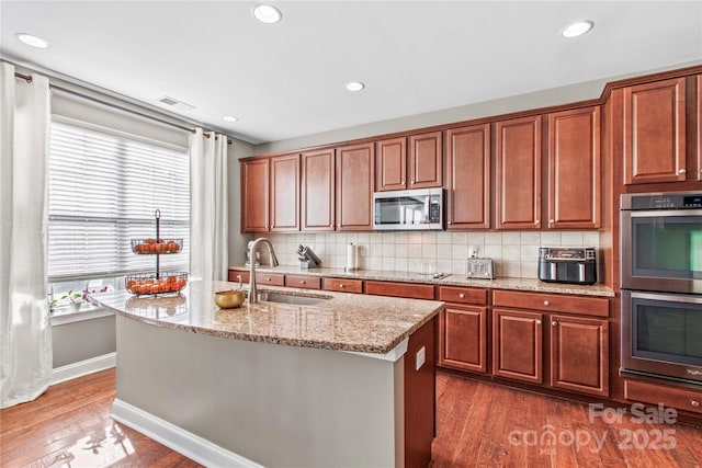 kitchen featuring sink, light stone counters, appliances with stainless steel finishes, a kitchen island with sink, and decorative backsplash