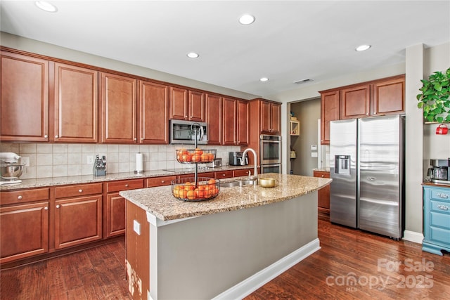 kitchen with sink, decorative backsplash, stainless steel appliances, light stone countertops, and a center island with sink