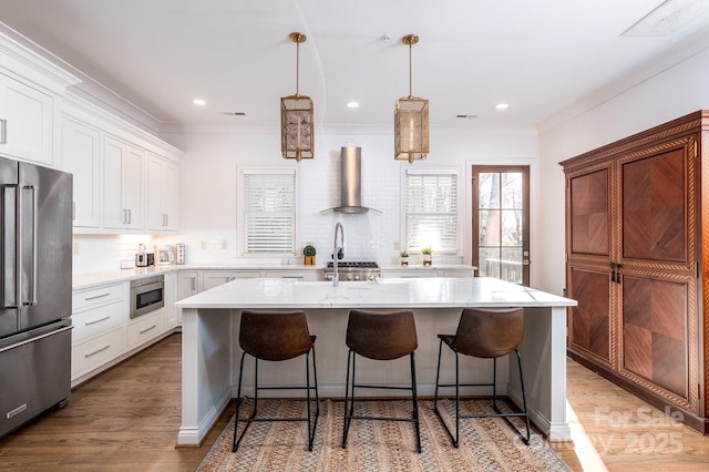 kitchen featuring white cabinets, an island with sink, wall chimney exhaust hood, hanging light fixtures, and stainless steel appliances