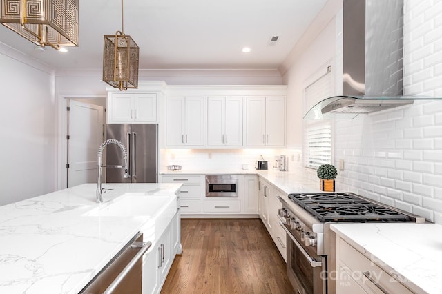 kitchen with premium appliances, wall chimney exhaust hood, light stone counters, hanging light fixtures, and white cabinetry