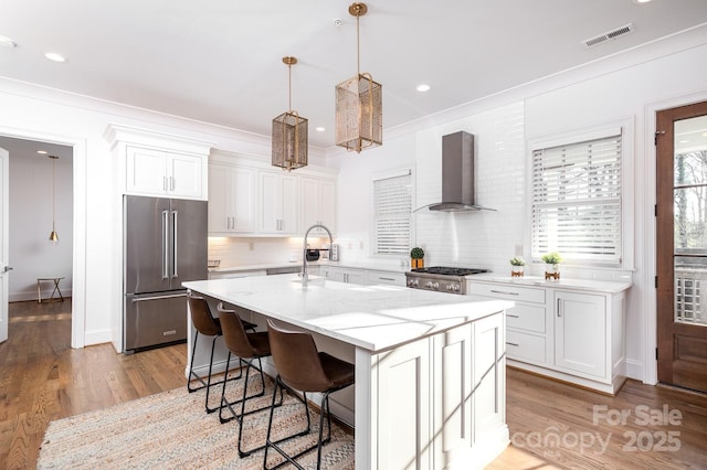 kitchen with high end fridge, wall chimney range hood, an island with sink, and white cabinets