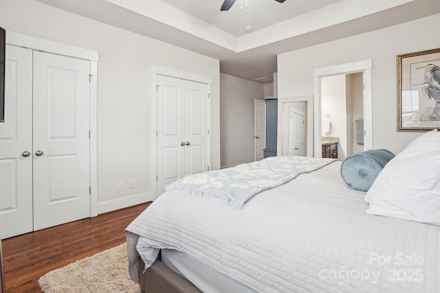 bedroom featuring ceiling fan, connected bathroom, a tray ceiling, dark hardwood / wood-style flooring, and multiple closets