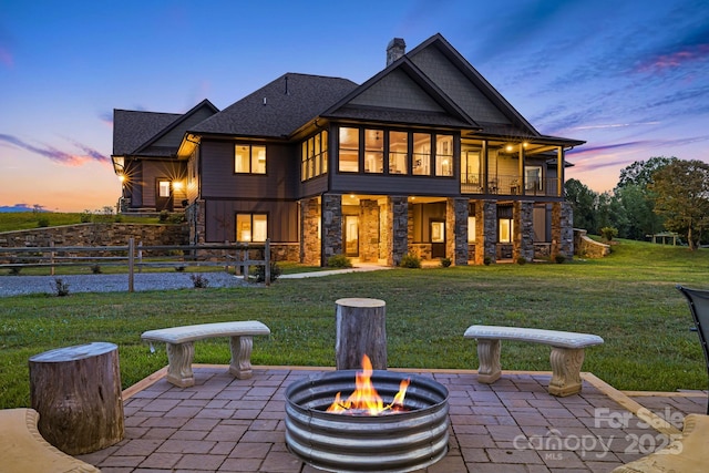 back house at dusk with a balcony, an outdoor fire pit, a patio, and a lawn