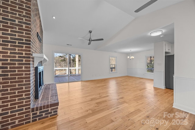 unfurnished living room featuring vaulted ceiling, a brick fireplace, ceiling fan with notable chandelier, and light wood-type flooring