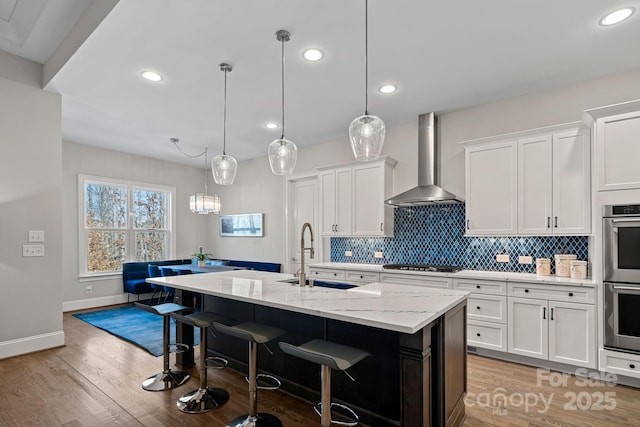 kitchen featuring sink, white cabinetry, decorative light fixtures, an island with sink, and wall chimney range hood