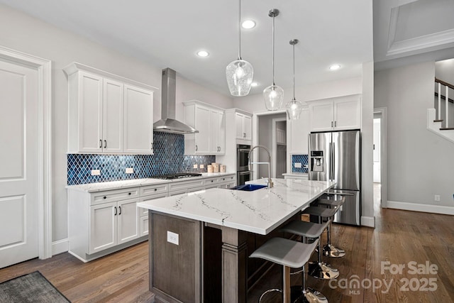 kitchen with white cabinetry, hanging light fixtures, a center island with sink, stainless steel appliances, and wall chimney range hood