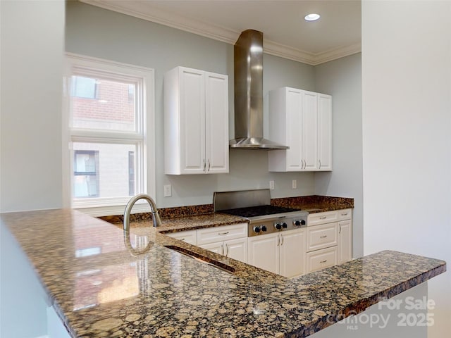 kitchen with crown molding, wall chimney range hood, kitchen peninsula, dark stone counters, and white cabinets