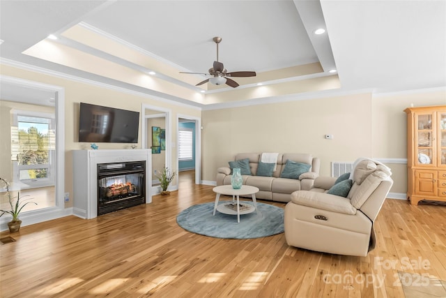 living room featuring crown molding, light wood-type flooring, a multi sided fireplace, a tray ceiling, and ceiling fan