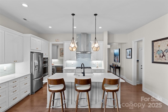 kitchen with appliances with stainless steel finishes, wall chimney exhaust hood, an island with sink, and hanging light fixtures