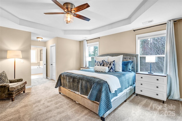 bedroom with ceiling fan, connected bathroom, a tray ceiling, and light carpet