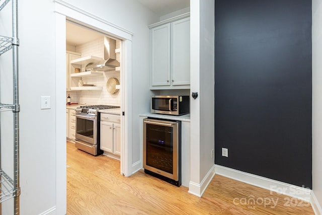 kitchen with appliances with stainless steel finishes, white cabinets, wine cooler, wall chimney exhaust hood, and light hardwood / wood-style flooring