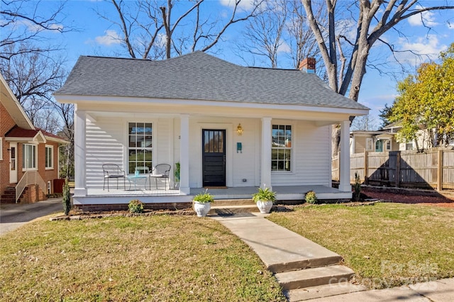 bungalow-style house featuring a shingled roof, a chimney, a porch, fence, and a front lawn