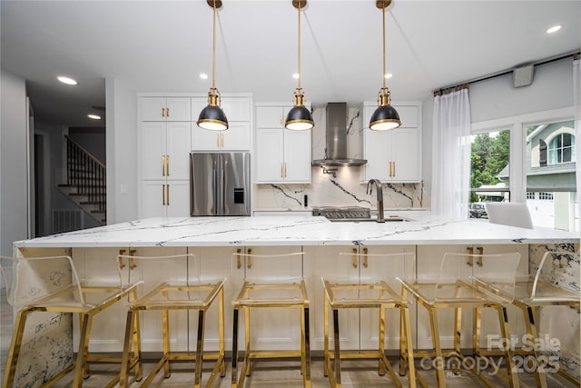 kitchen with light stone counters, wall chimney range hood, stainless steel fridge, and white cabinets