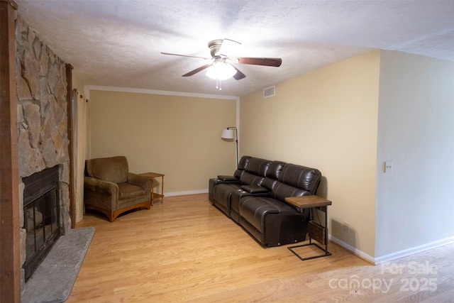 living room with ceiling fan, a stone fireplace, a textured ceiling, and light wood-type flooring