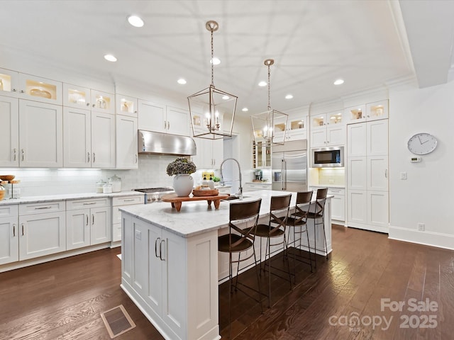 kitchen with built in appliances, pendant lighting, a kitchen island with sink, and white cabinets