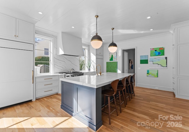 kitchen featuring wall chimney range hood, a breakfast bar, paneled built in fridge, an island with sink, and white cabinets