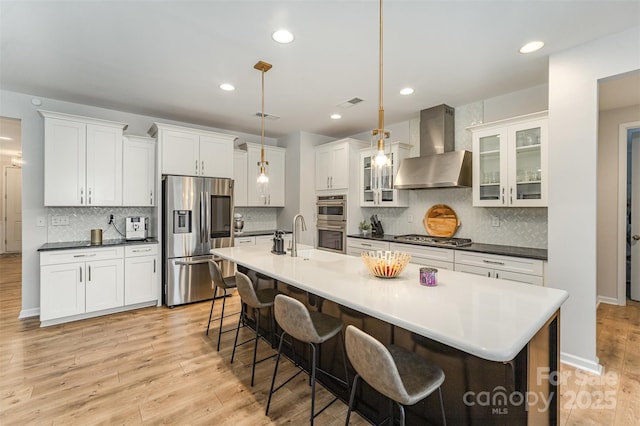 kitchen with a breakfast bar, appliances with stainless steel finishes, a kitchen island with sink, decorative light fixtures, and wall chimney exhaust hood