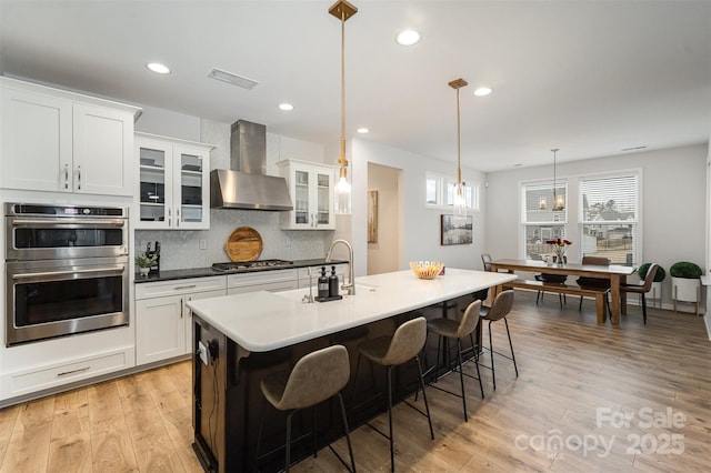 kitchen featuring hanging light fixtures, stainless steel appliances, white cabinets, a kitchen bar, and wall chimney exhaust hood