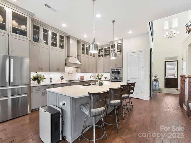 kitchen featuring pendant lighting, gray cabinets, appliances with stainless steel finishes, a center island with sink, and custom exhaust hood