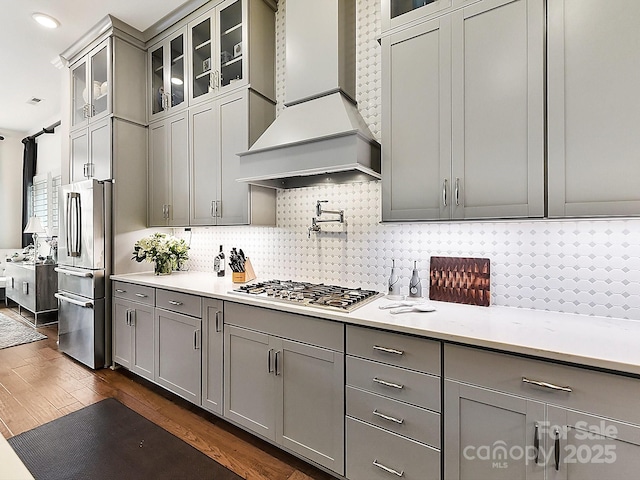 kitchen with dark wood-type flooring, gray cabinets, premium range hood, appliances with stainless steel finishes, and tasteful backsplash