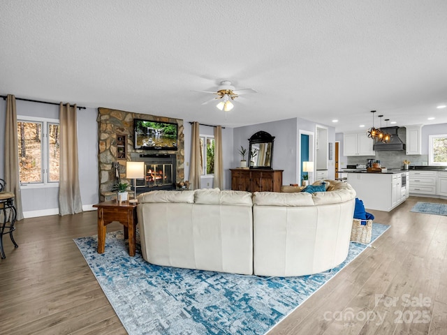 living room with ceiling fan, a stone fireplace, light hardwood / wood-style flooring, and a textured ceiling