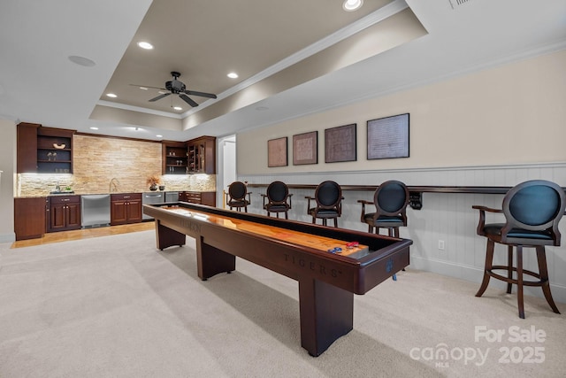 recreation room featuring a raised ceiling, ornamental molding, light carpet, and indoor wet bar