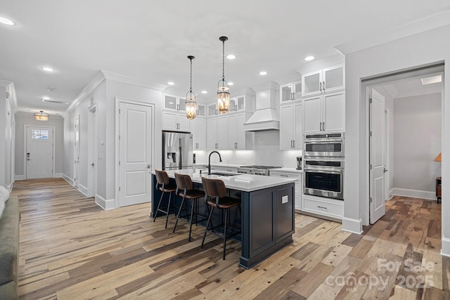 kitchen featuring premium range hood, appliances with stainless steel finishes, white cabinetry, an island with sink, and sink