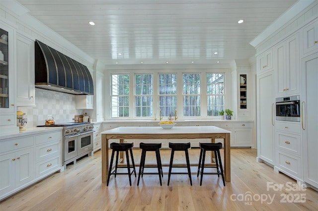 kitchen with glass insert cabinets, light countertops, stainless steel appliances, wall chimney range hood, and white cabinets