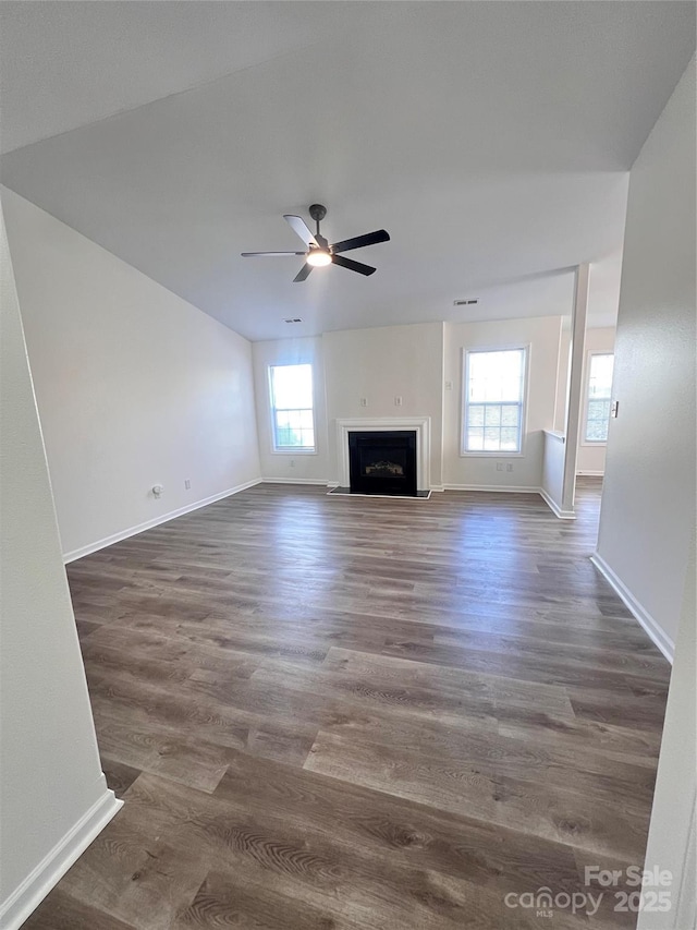 unfurnished living room with a wealth of natural light, a ceiling fan, a fireplace with raised hearth, and dark wood-type flooring