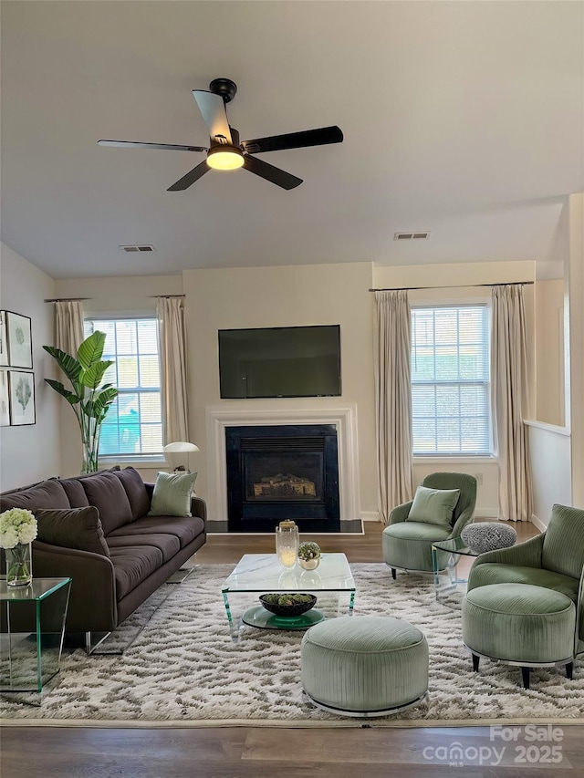 living room featuring a ceiling fan, a glass covered fireplace, visible vents, and wood finished floors