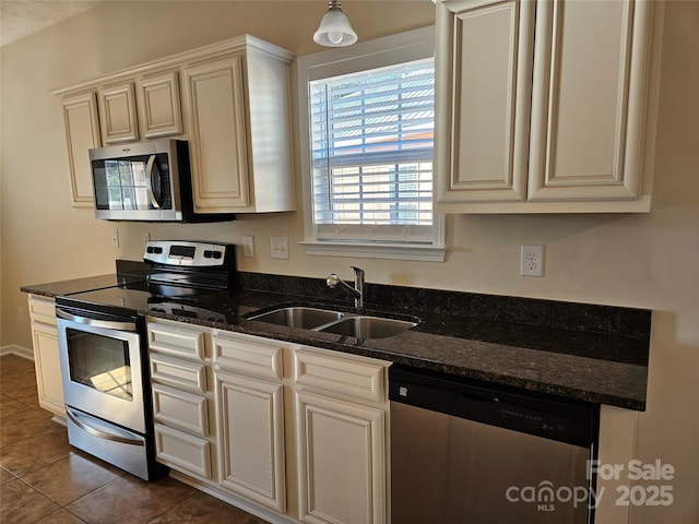 kitchen with stainless steel appliances, a sink, cream cabinetry, dark stone countertops, and pendant lighting