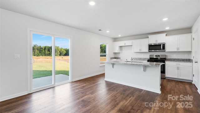 kitchen with dark hardwood / wood-style floors, white cabinets, stainless steel appliances, light stone countertops, and a center island with sink